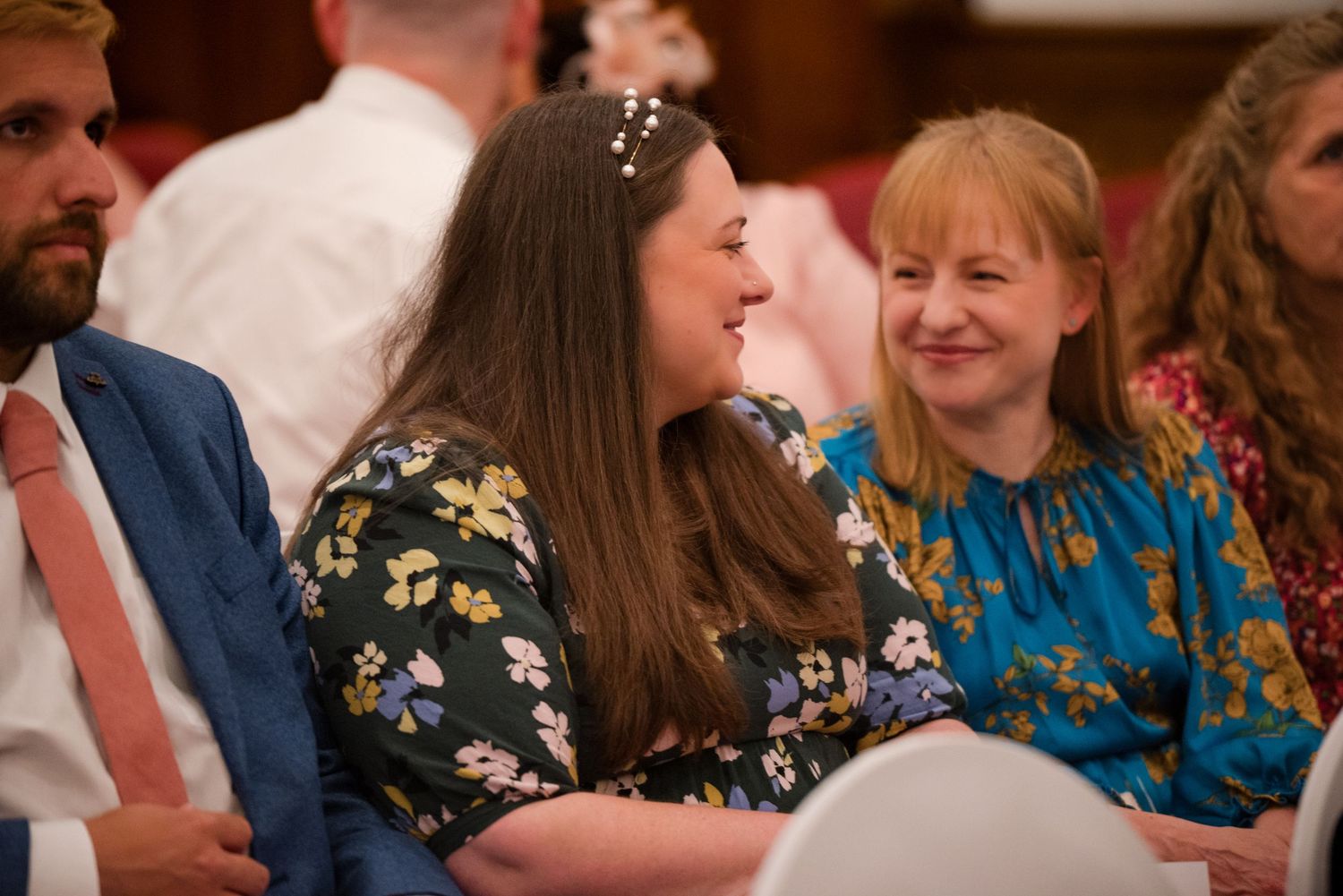 Wedding guests seated in rows smile and laugh during a reception celebration.