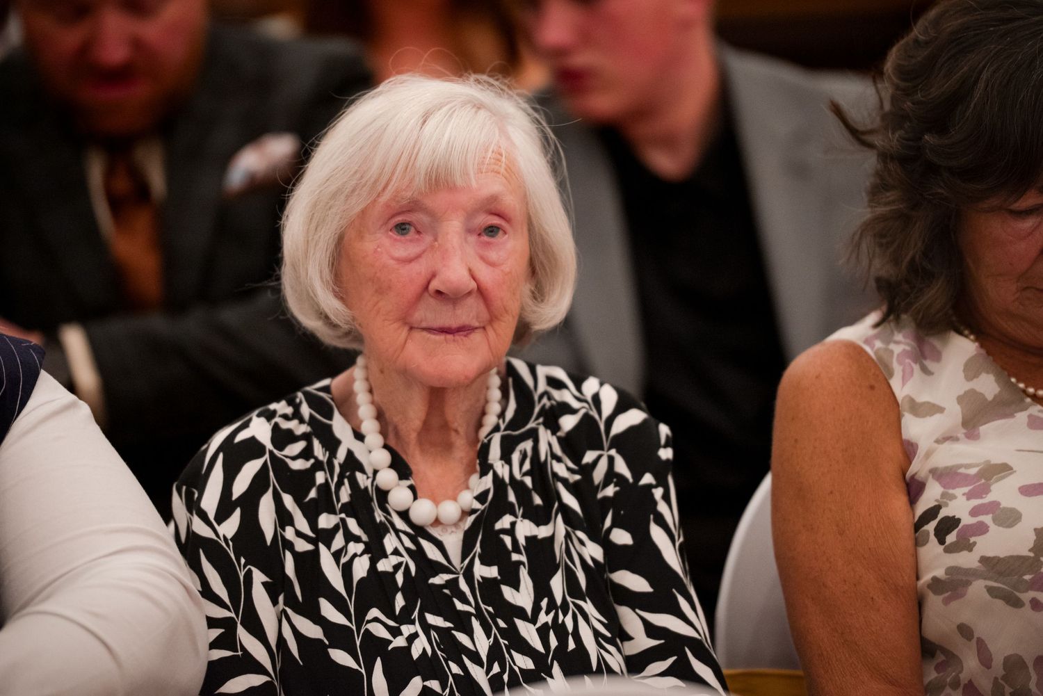 A person with white hair wearing a black and white patterned outfit smiles while seated at a social gathering.