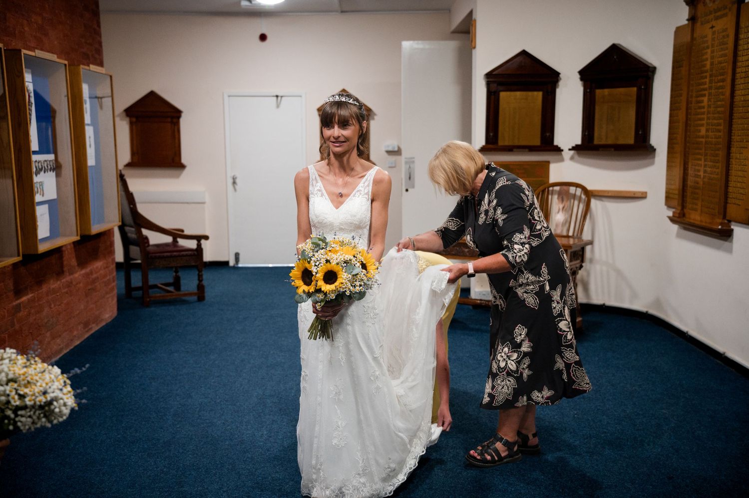A person in a white wedding dress holds a sunflower bouquet while someone assists with the dress in a blue-carpeted room.