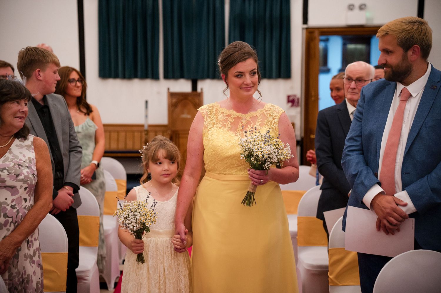Wedding guests in formal attire gather at a reception, with yellow bridesmaid dresses and baby's breath bouquets visible.