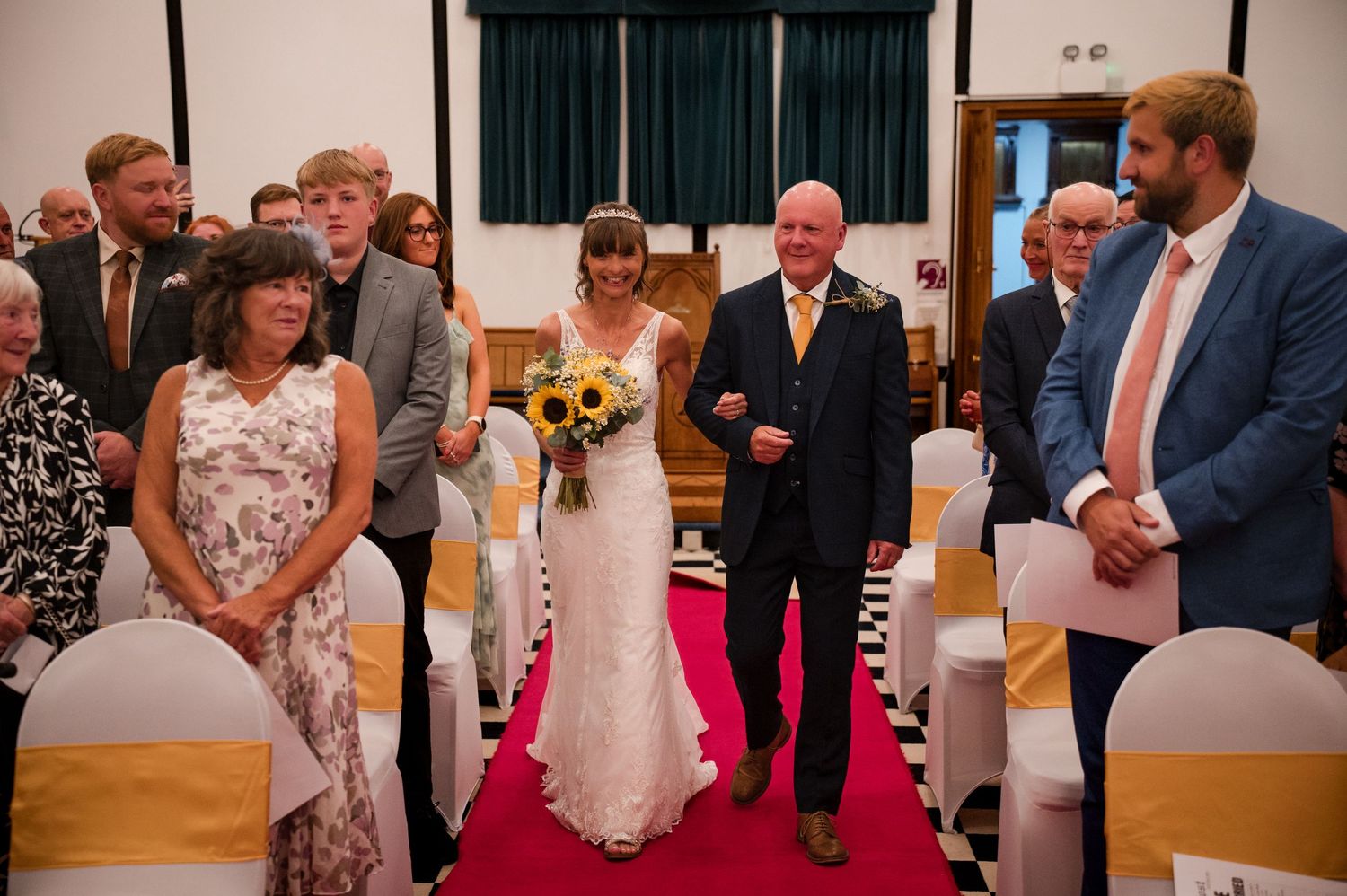 Wedding guests watching a ceremony in a church hall with wooden floors and red carpet.