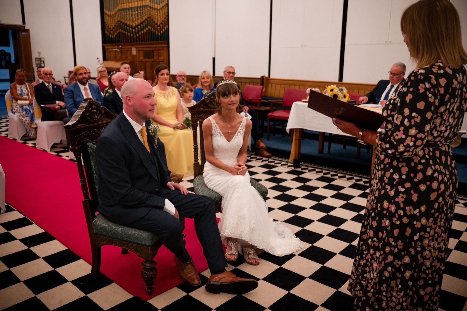 Wedding guests seated in rows of chairs on a black and white checkered floor during an indoor ceremony.