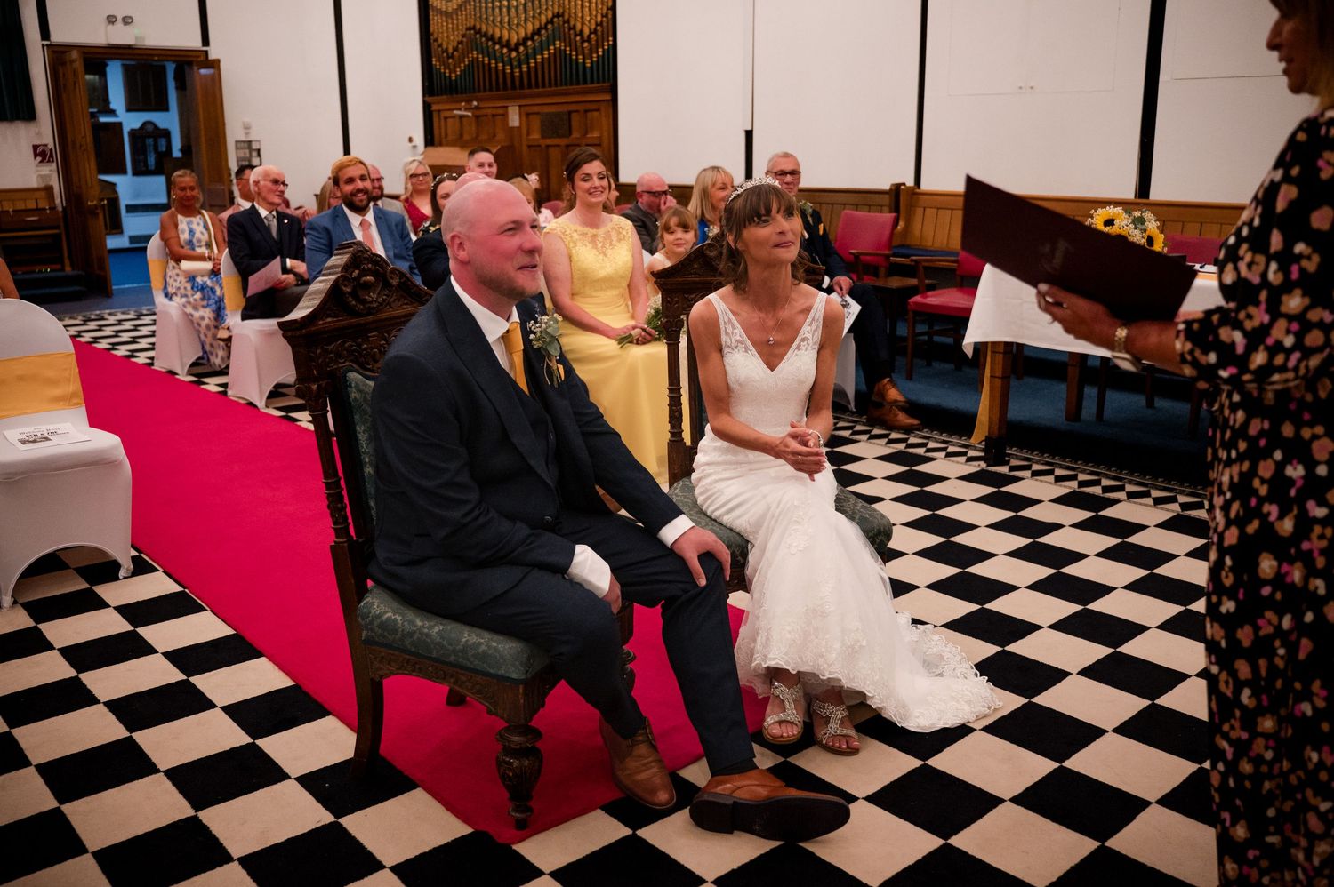 Wedding guests sit in chairs on a black and white checkered floor during an indoor ceremony.