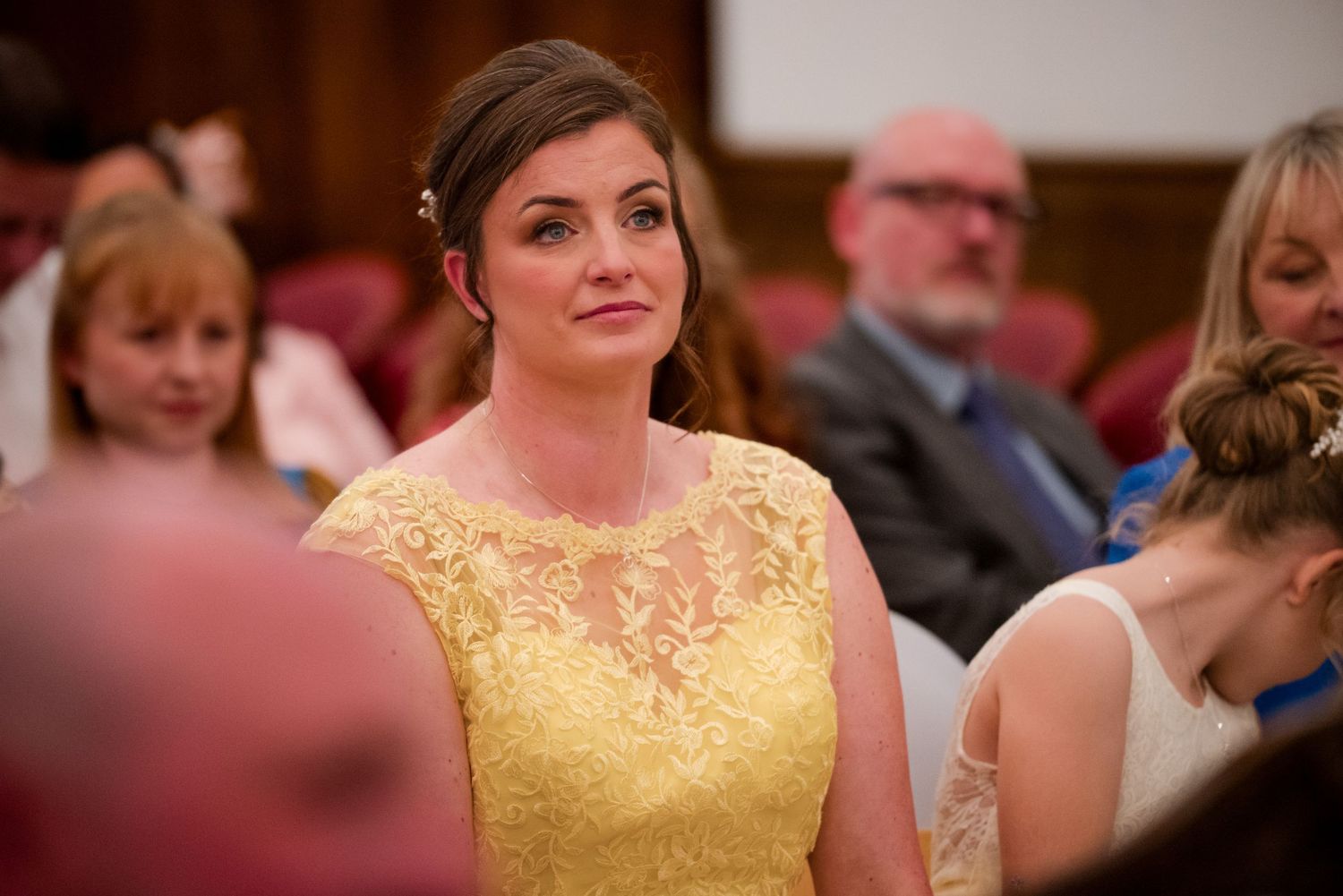 A person in a bright yellow lace dress is seen in focus at what appears to be a formal event or gathering.