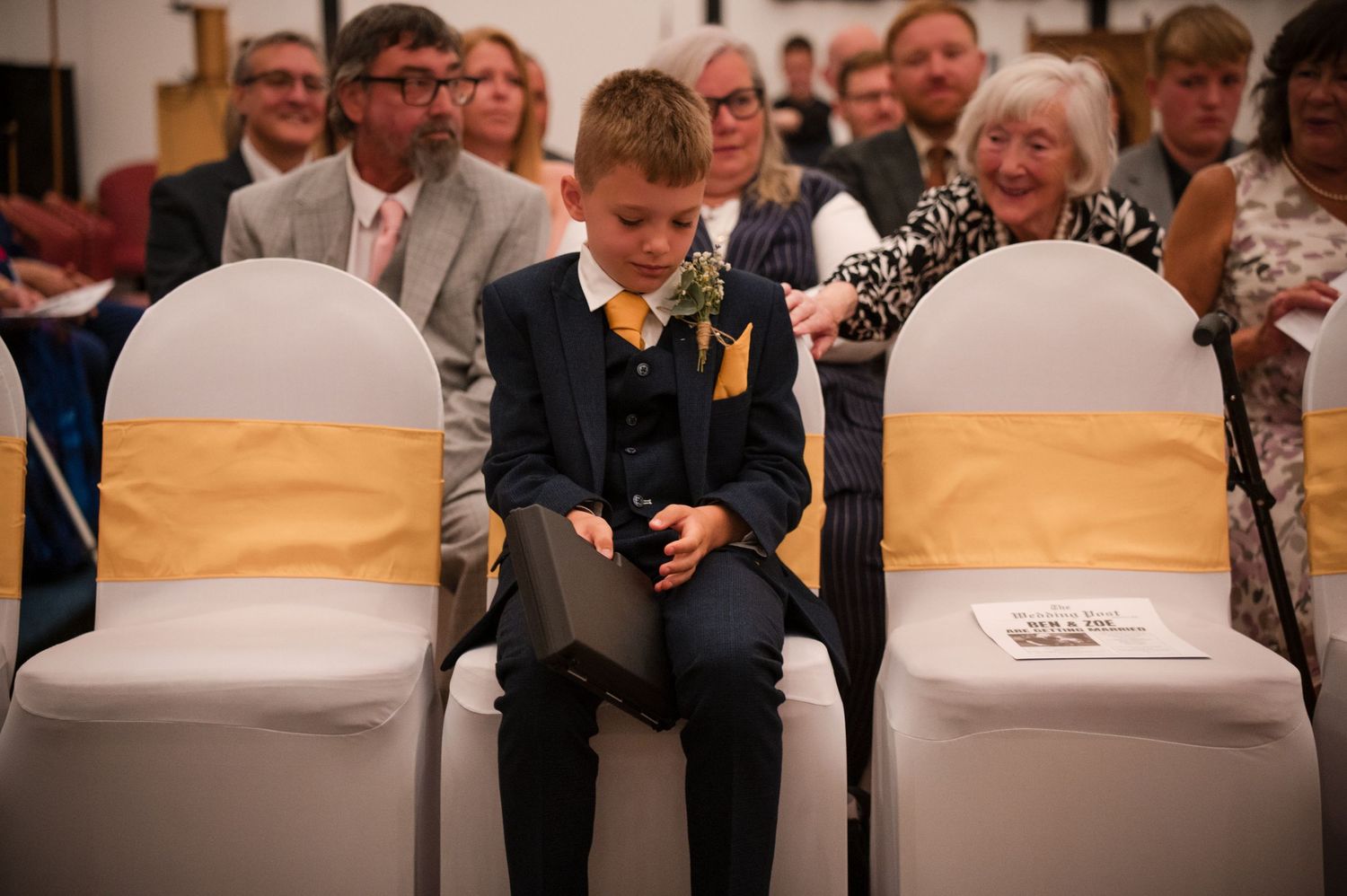 A young attendee in a dark suit sits between white chairs with gold sashes at a formal event.