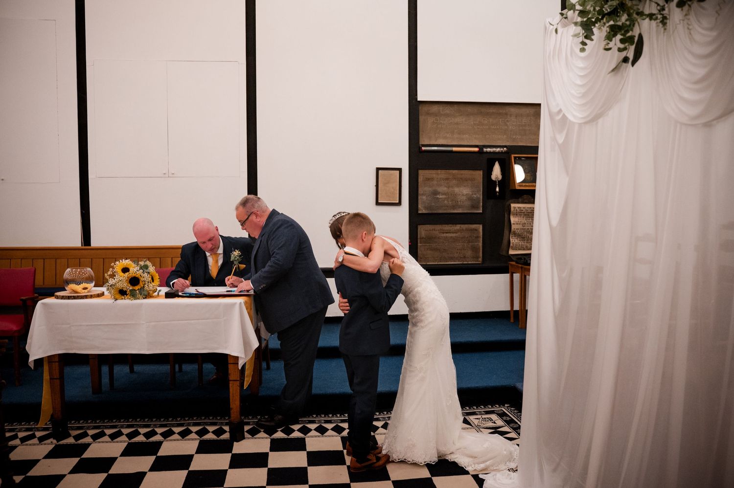 Two people in formal attire share a moment at a dining table with a black and white checkered floor.