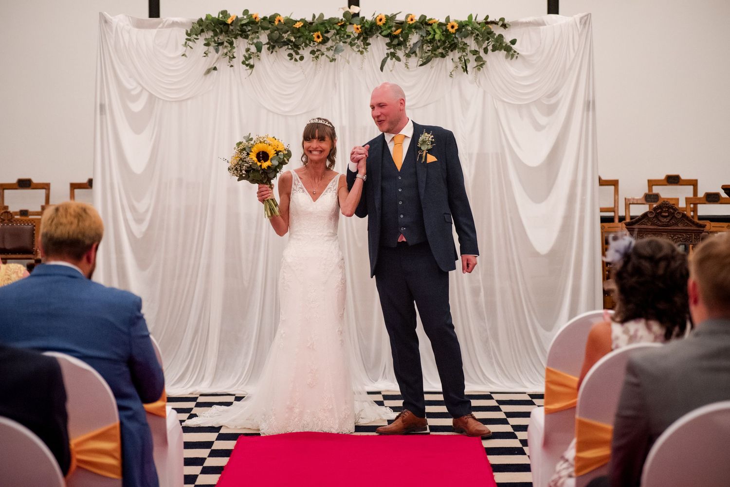 A wedding ceremony takes place in front of white draped fabric backdrop decorated with greenery and floral arrangements.