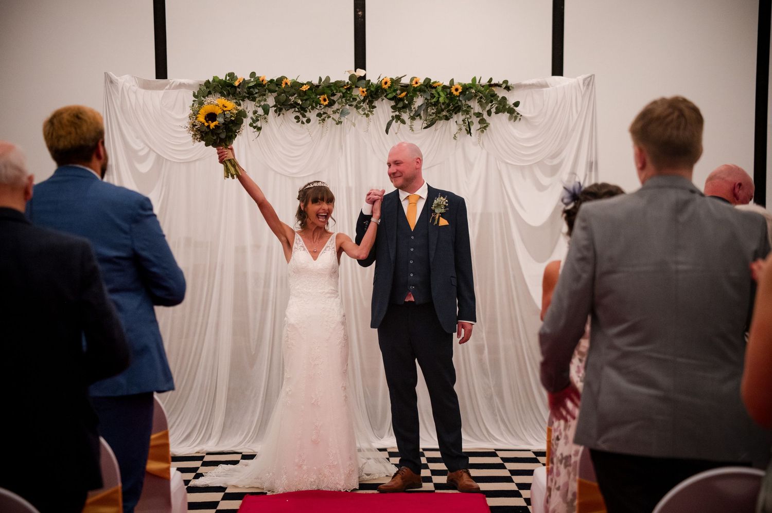 Wedding guests dance and celebrate on a checkered dance floor under a white tent at an evening reception.