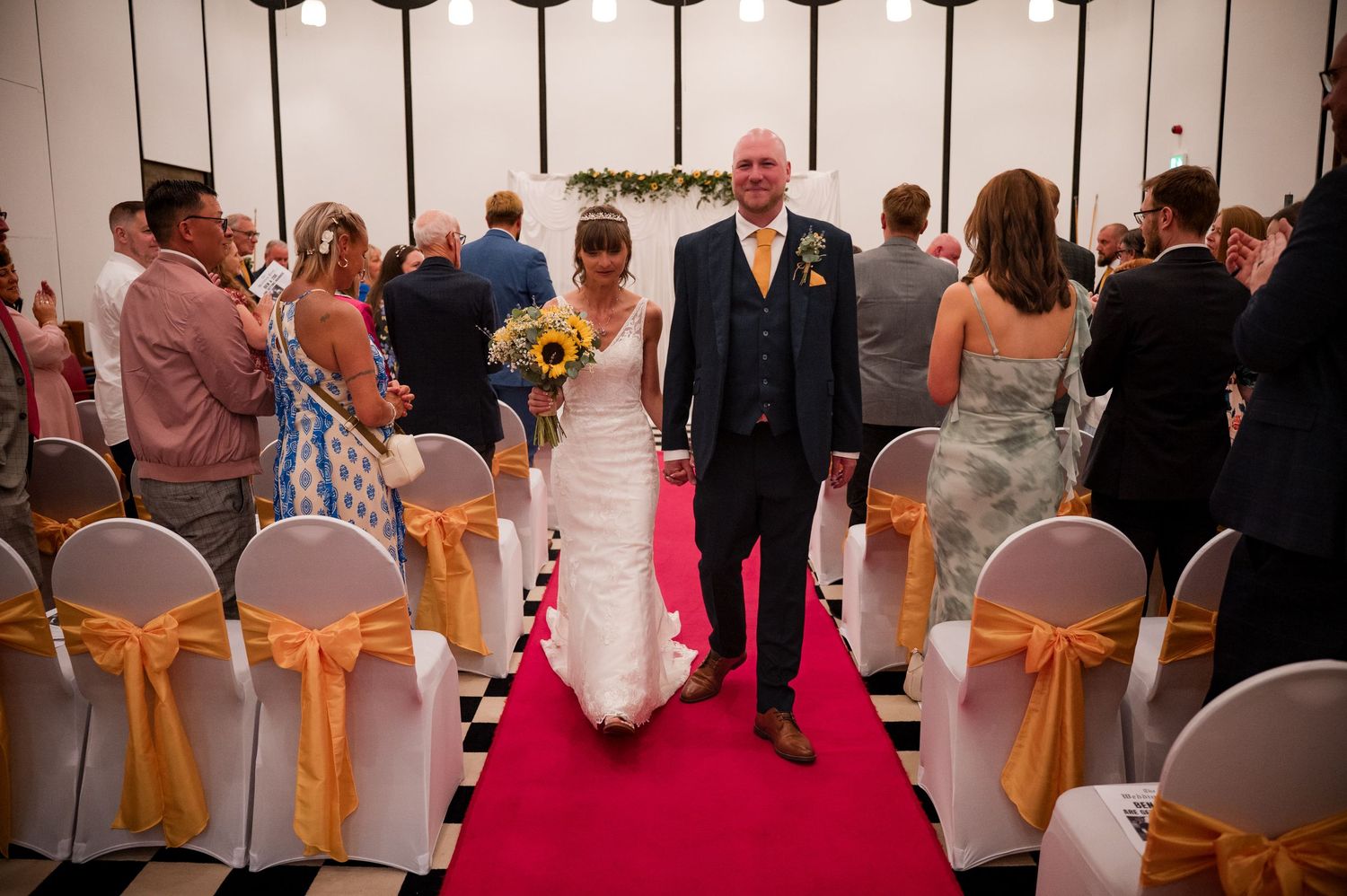 A wedding reception venue with chairs decorated in gold sashes and guests mingling in the background.