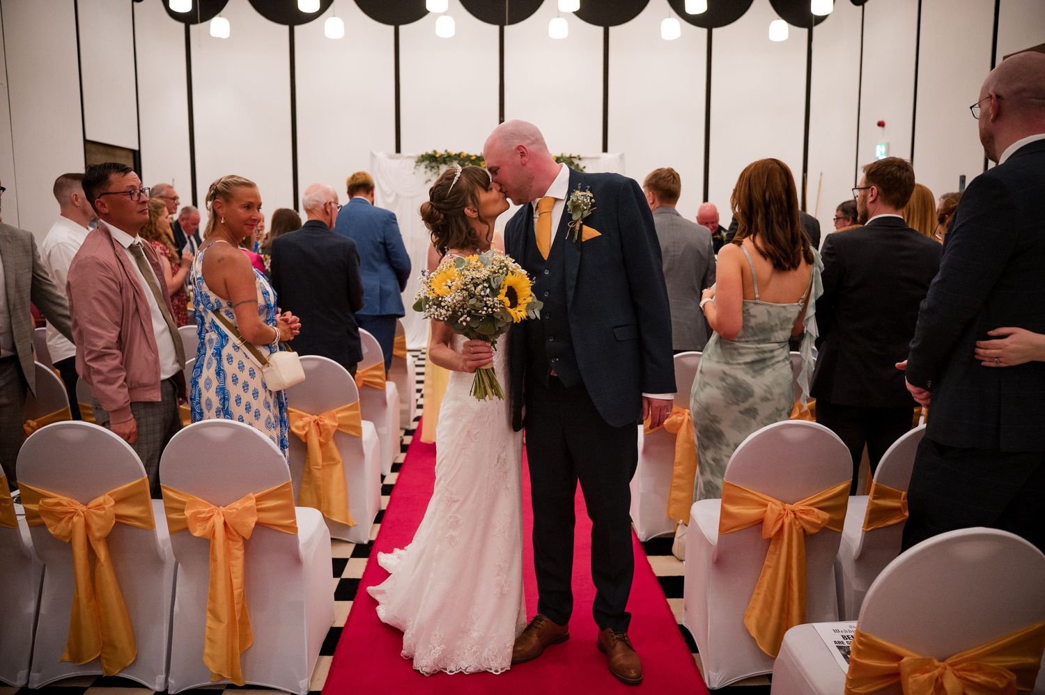 Wedding guests dance and celebrate during a reception in a banquet hall decorated with orange chair sashes.