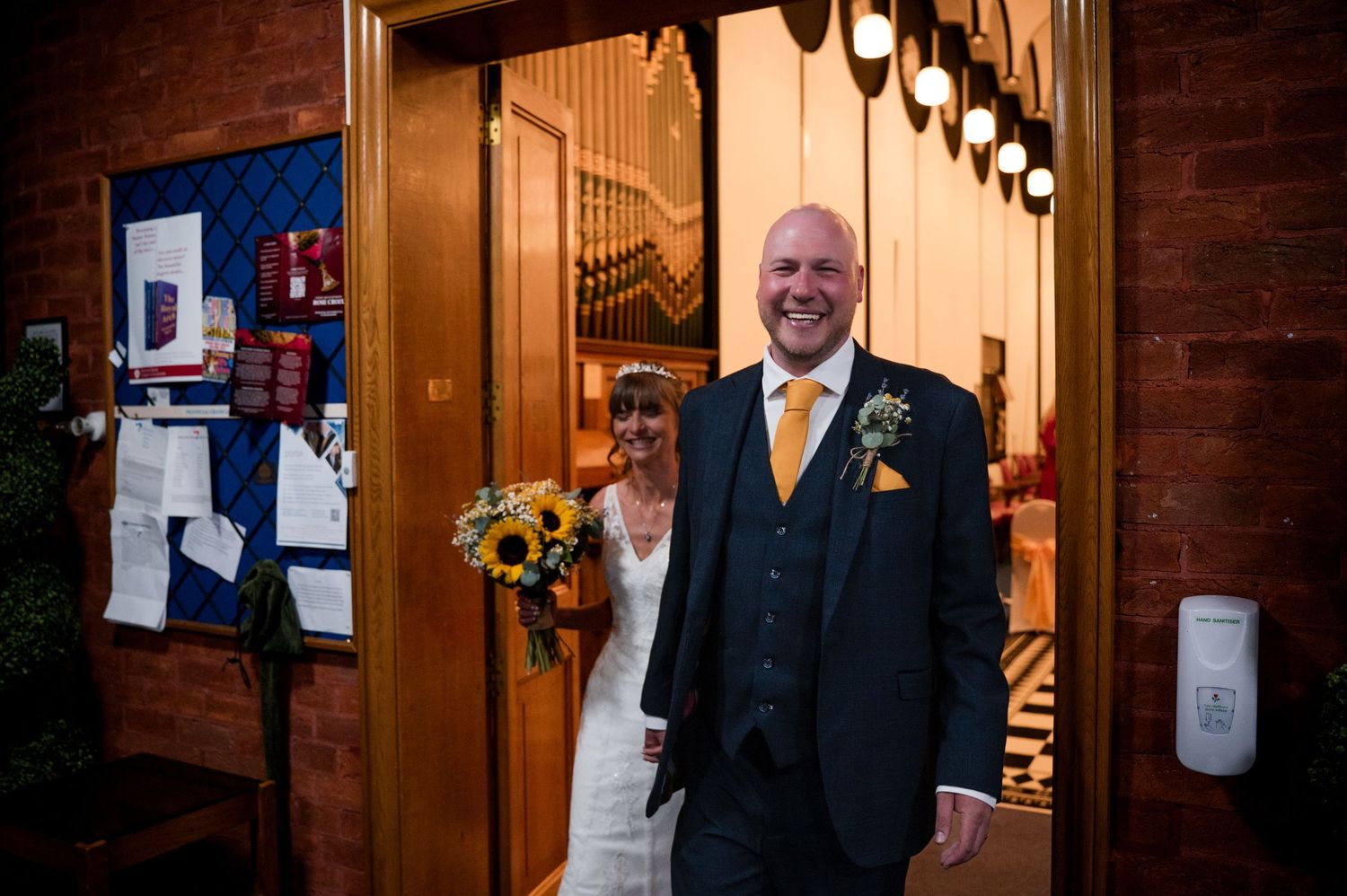 A person in a black suit and orange tie stands in a wood-paneled hallway with pendant lights overhead.
