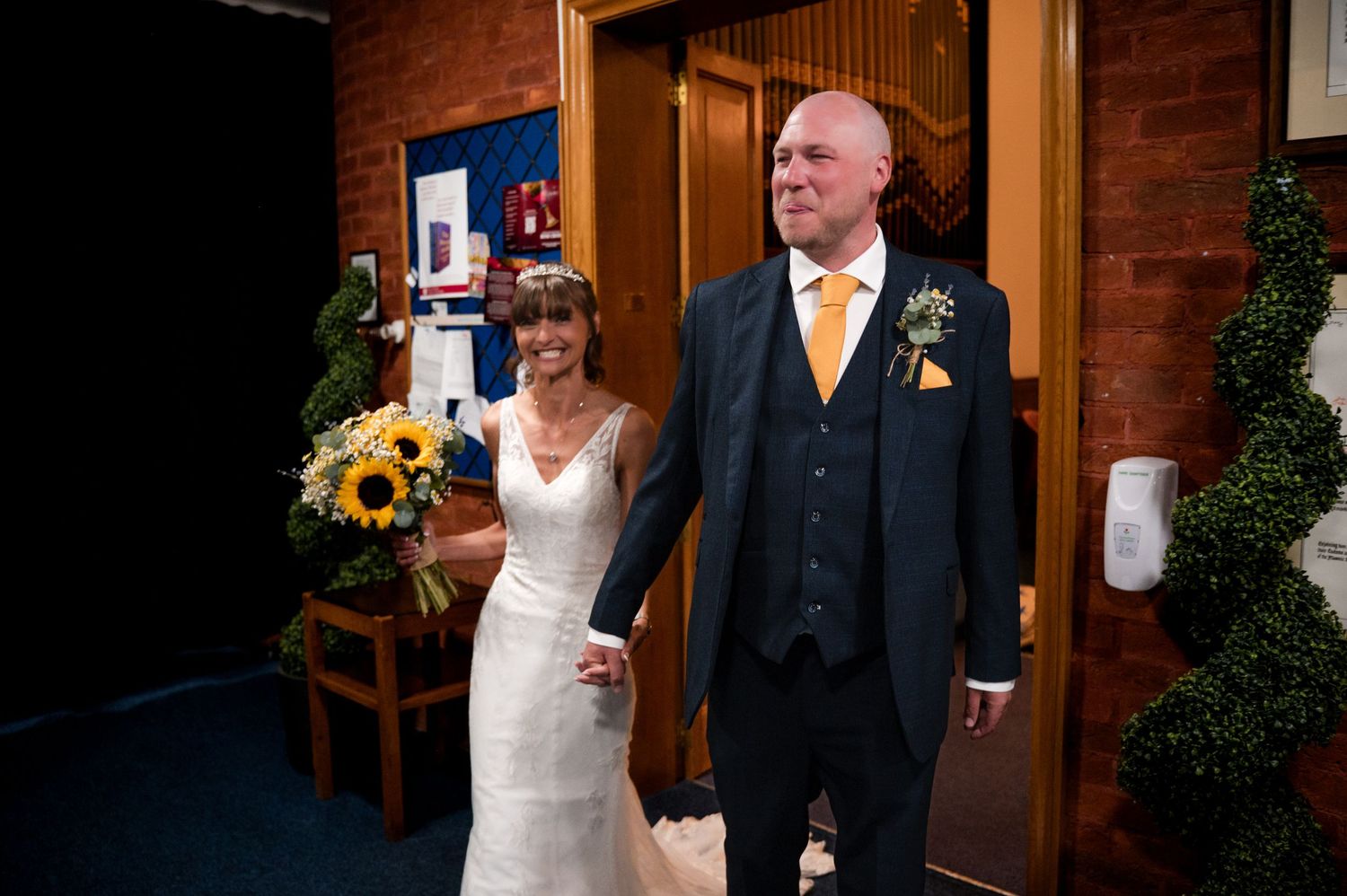 A couple in formal wedding attire hold hands while standing in a church entryway decorated with sunflowers.
