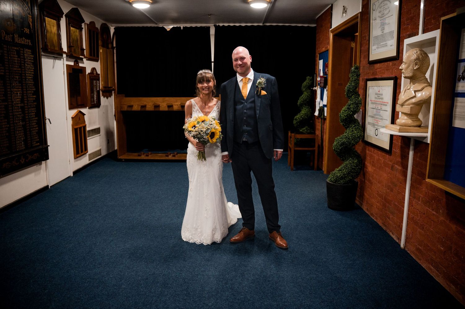A couple in formal wedding attire walks together down a hallway with wooden walls and blue carpeting.