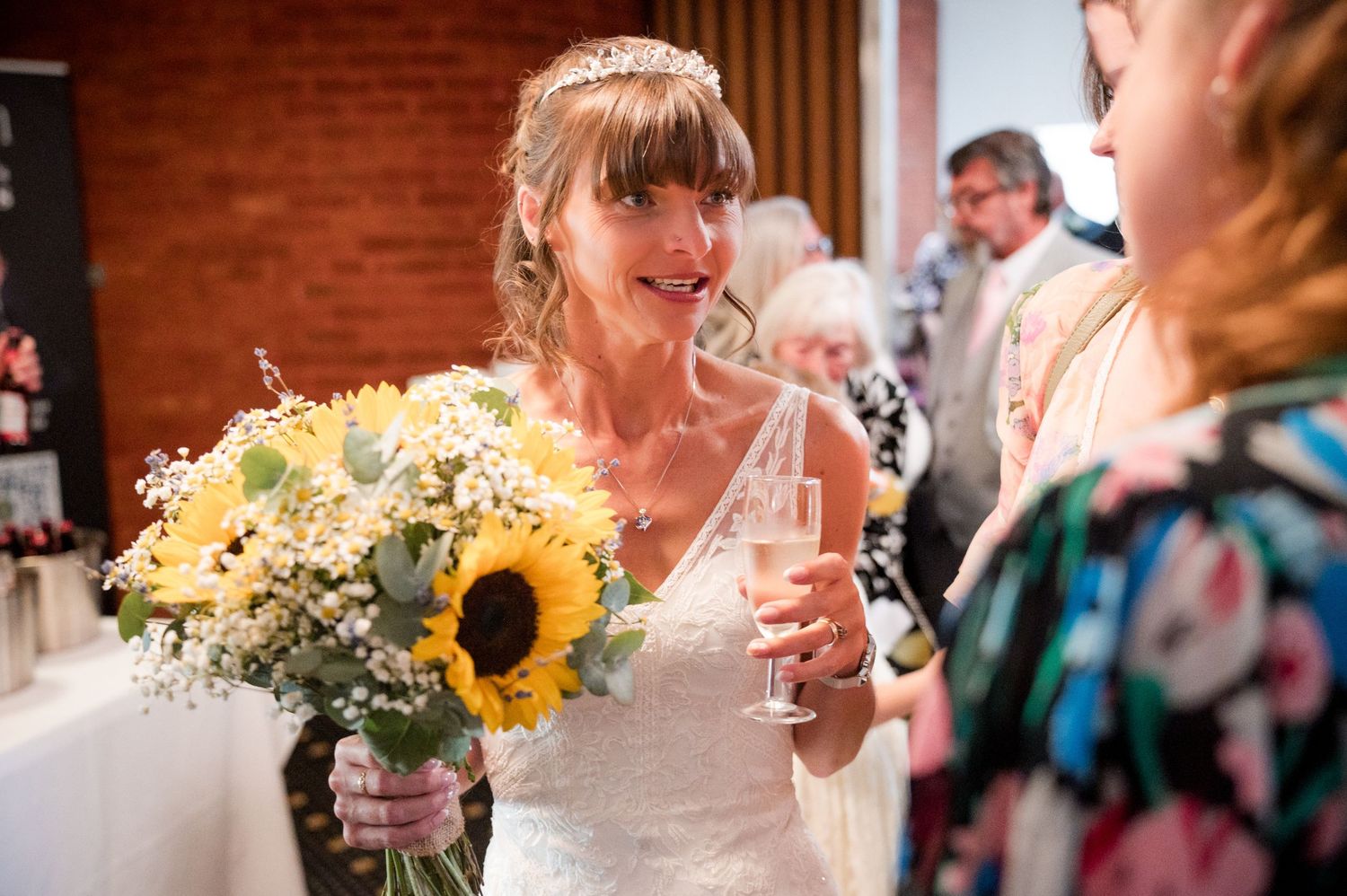 A bride in a white dress holds a bright sunflower bouquet while enjoying a drink at a wedding reception.