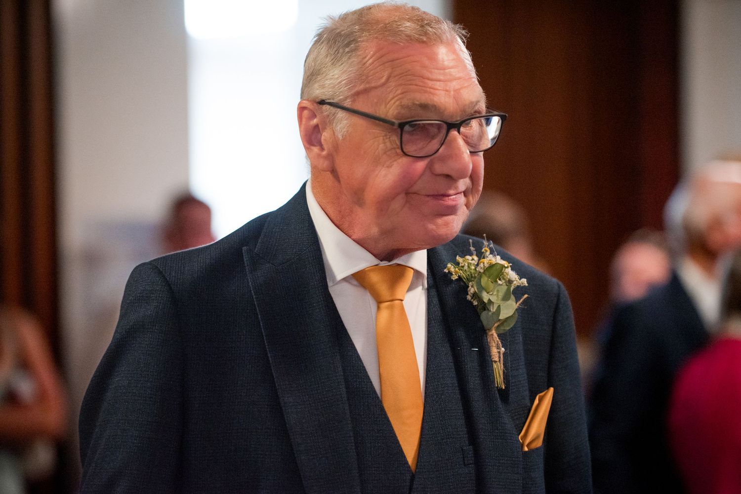 A well-dressed guest at a formal event wearing a navy suit with gold tie and boutonniere.