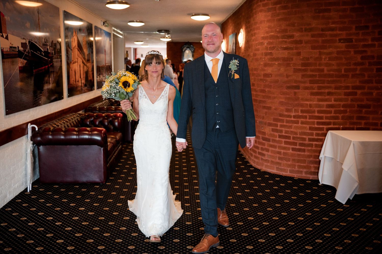 Wedding procession walking through an indoor venue with exposed brick walls and dark patterned carpeting.
