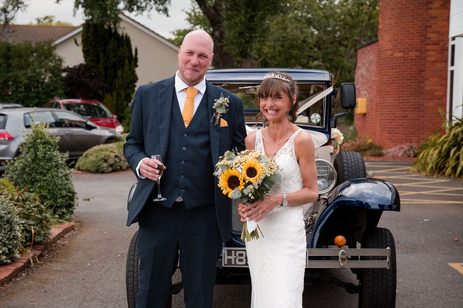 Wedding couple poses with sunflower bouquet next to vintage black car outside brick building.