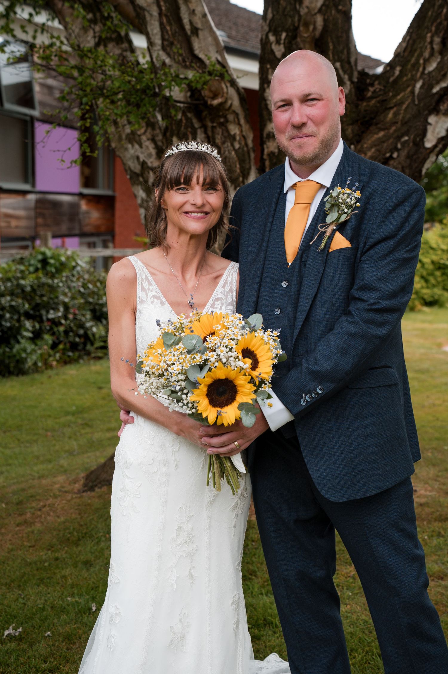 Wedding couple poses outdoors with bright yellow sunflower bouquet and navy suit against garden backdrop.