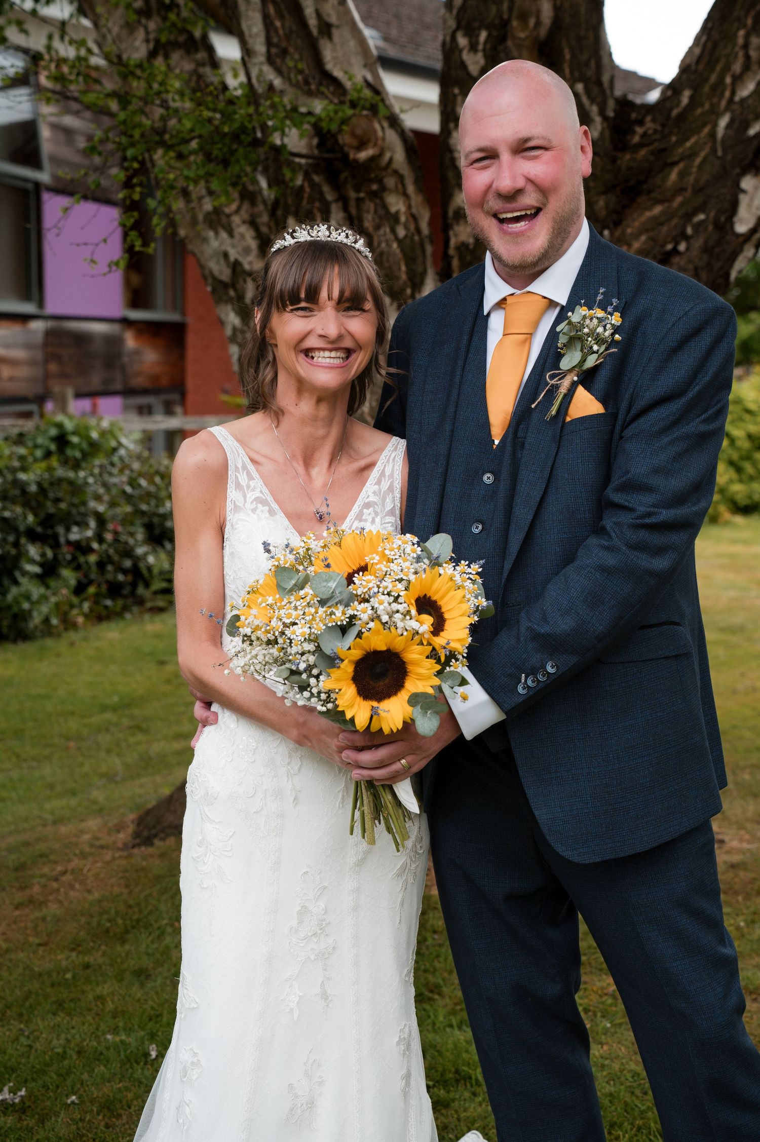 Wedding couple poses outdoors with bright sunflower bouquet and navy suit with orange tie accents.