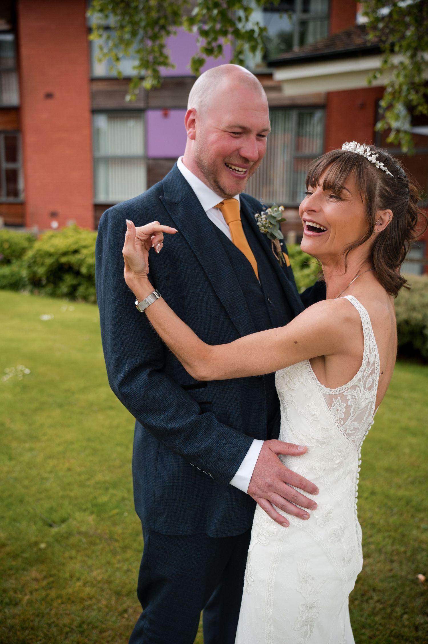 A joyful wedding couple shares a candid laughing moment together on a lawn outside a brick building.
