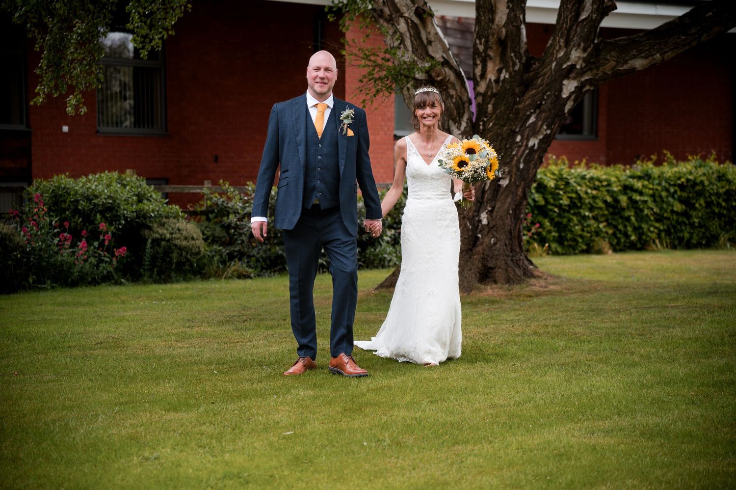 A couple in wedding attire stands together on a lush green lawn near a red brick building.