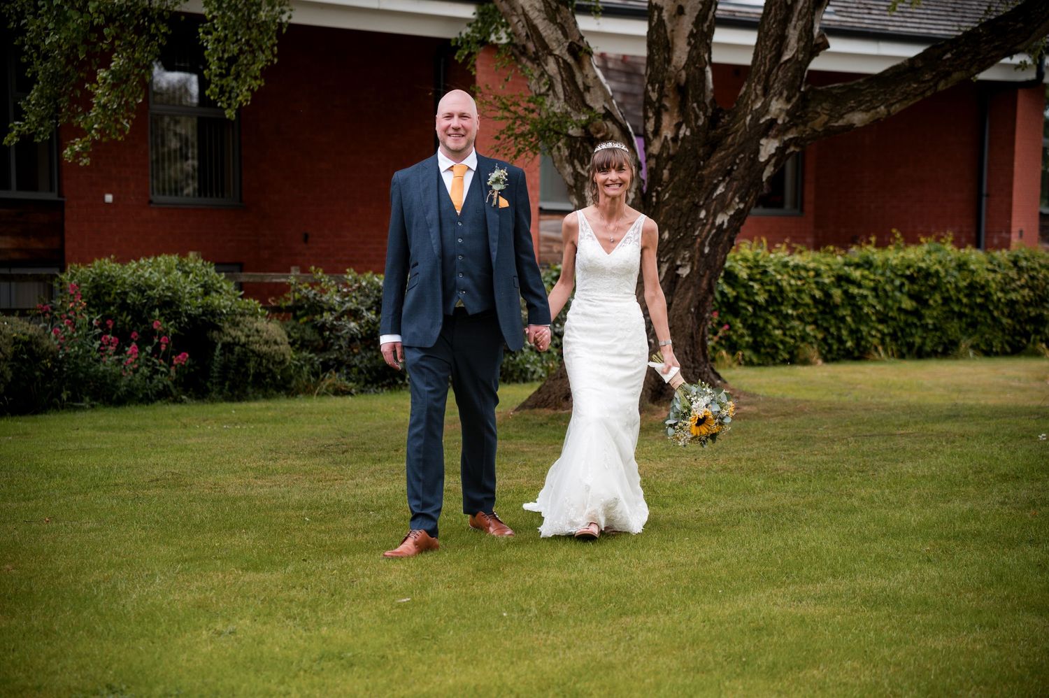 Newlyweds walk and laugh together on a lush green lawn outside a red brick building during their wedding celebration.