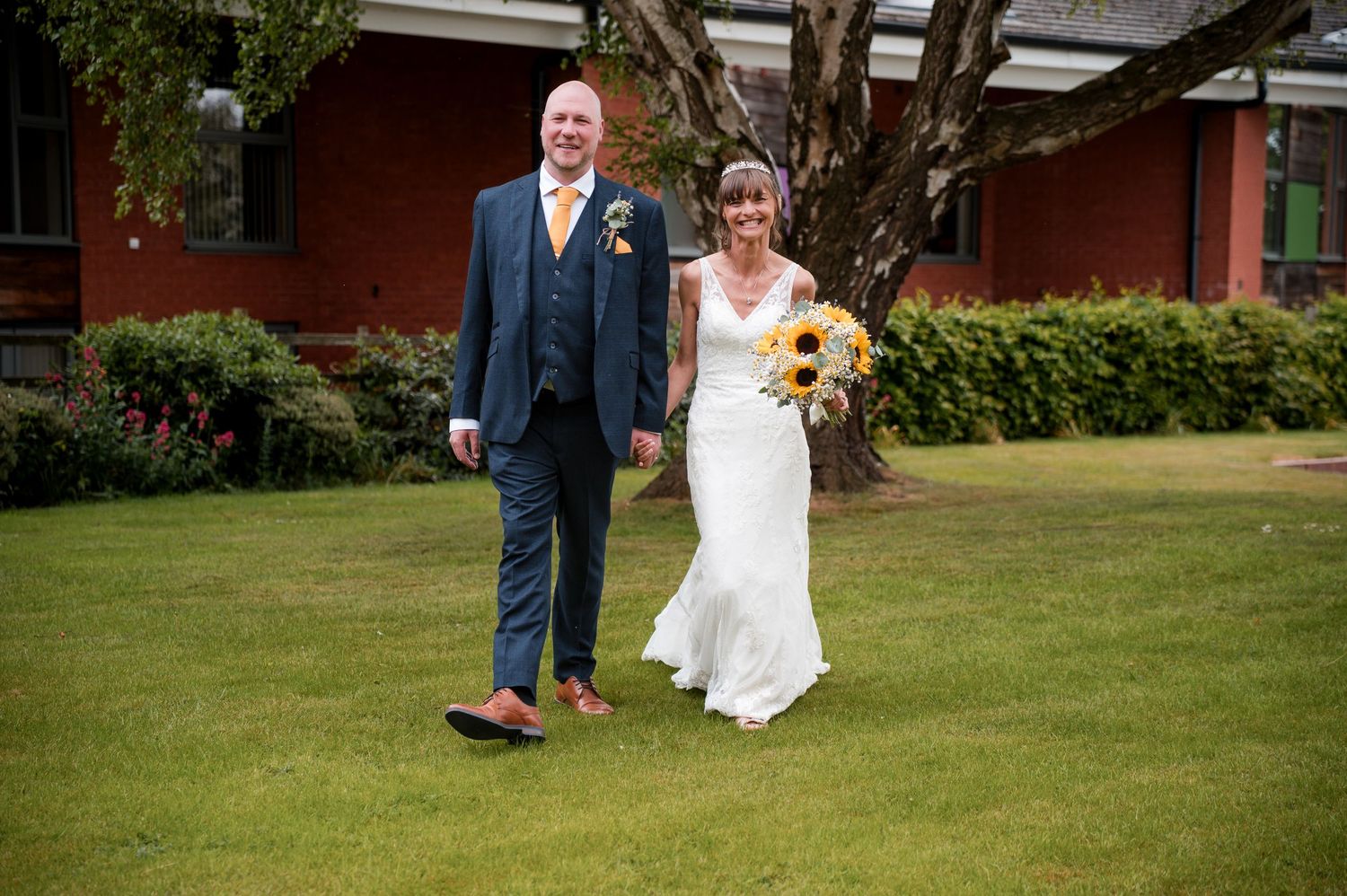 Wedding guests celebrate by throwing confetti over a couple walking on a lush green lawn by a brick building.