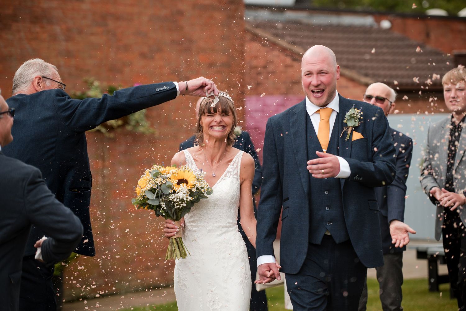 Wedding guests toss confetti over newlyweds outside a brick building during a joyful celebration.