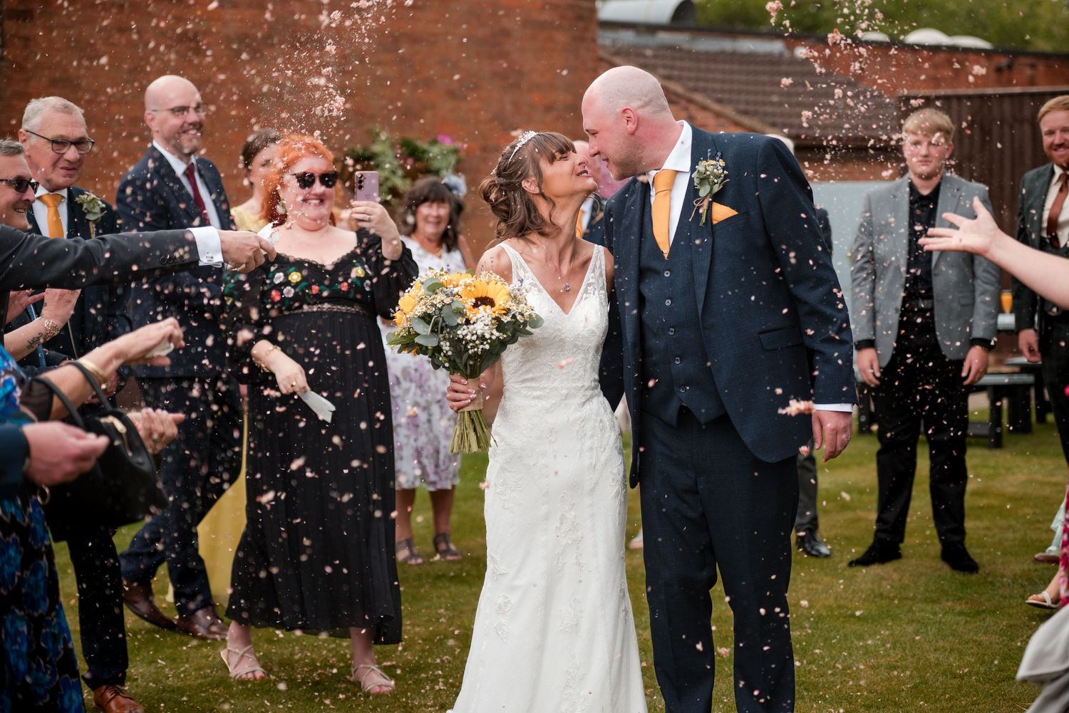 Wedding guests shower the newlyweds with confetti outside a rustic brick venue during a joyful celebration.