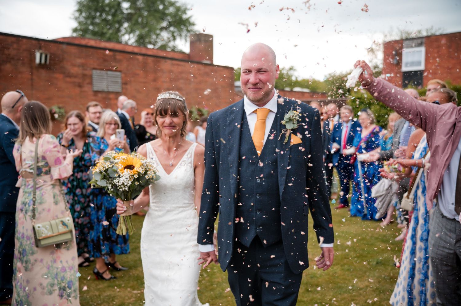 Wedding guests throw confetti at a joyful celebration outside a brick building on a sunny day.