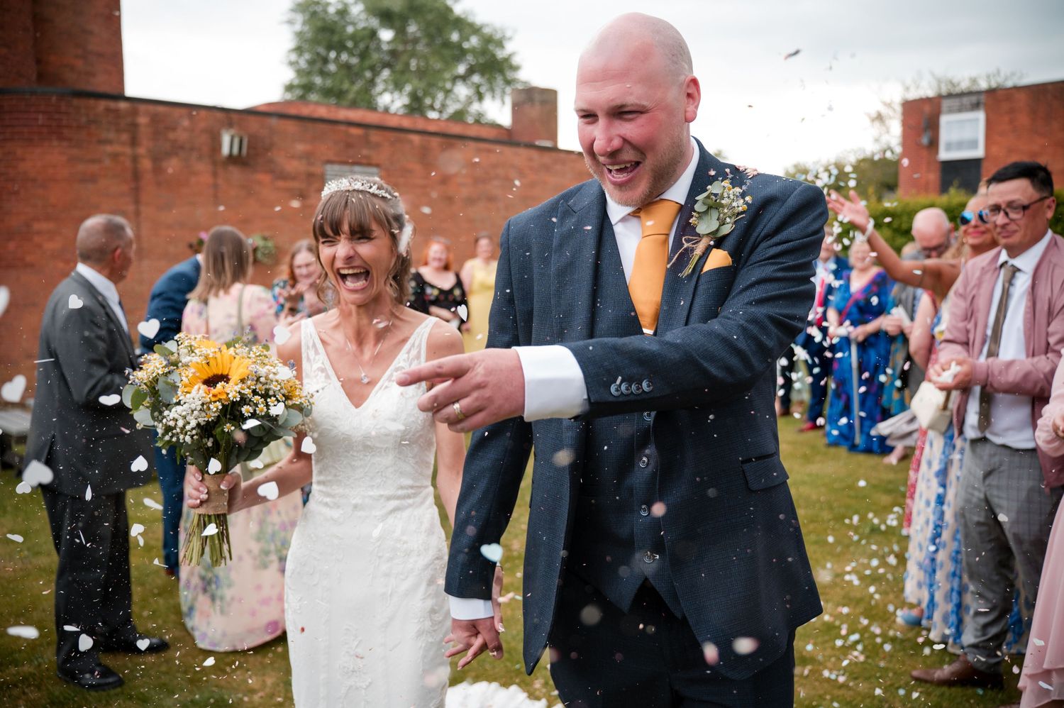 Wedding guests celebrate with confetti toss outside red brick building during joyful reception moment.