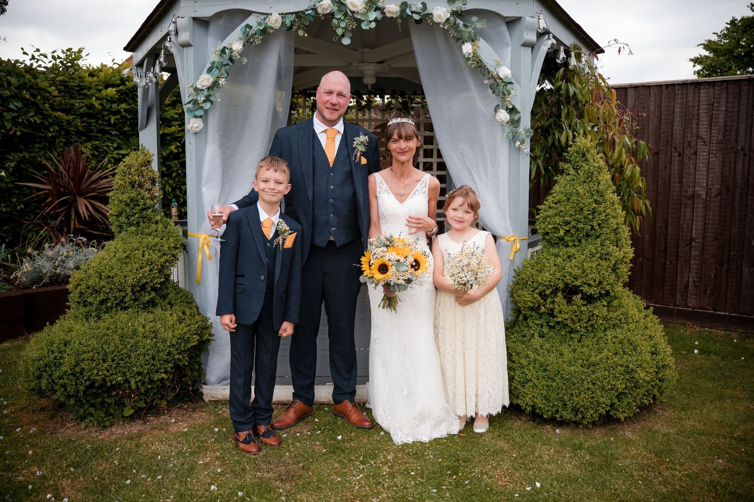 A wedding party poses under a white draped garden arch with colorful floral bouquets in a backyard setting.