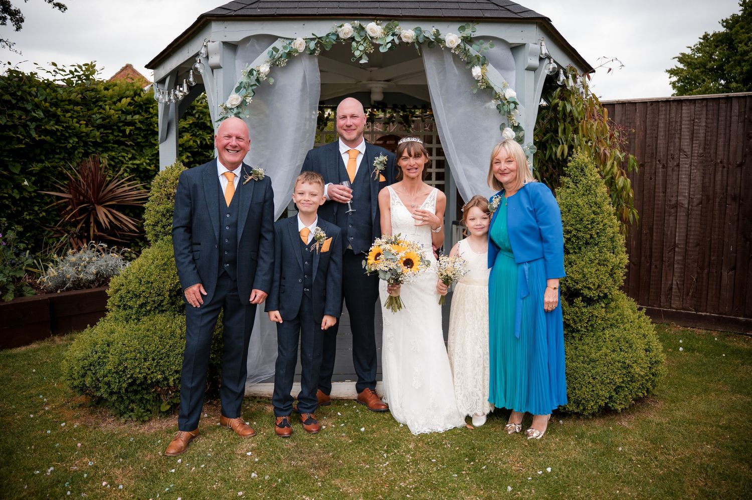 A group stands together for a formal photo in front of a white garden gazebo surrounded by greenery.