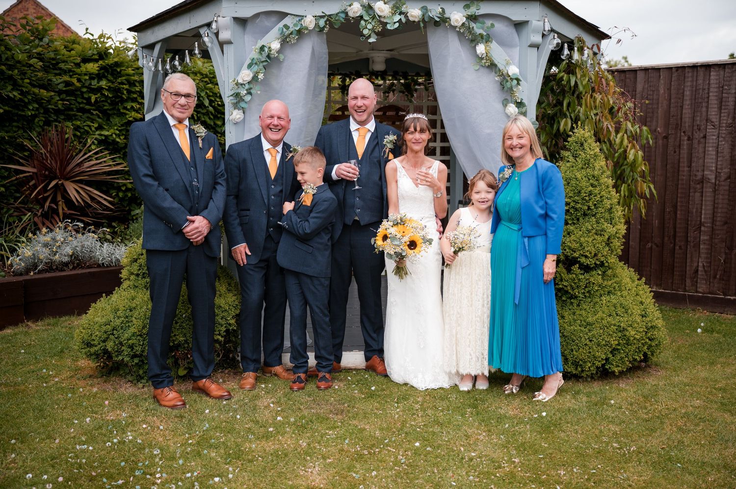 Wedding party poses together outdoors in garden setting with decorative floral arrangements and greenery.