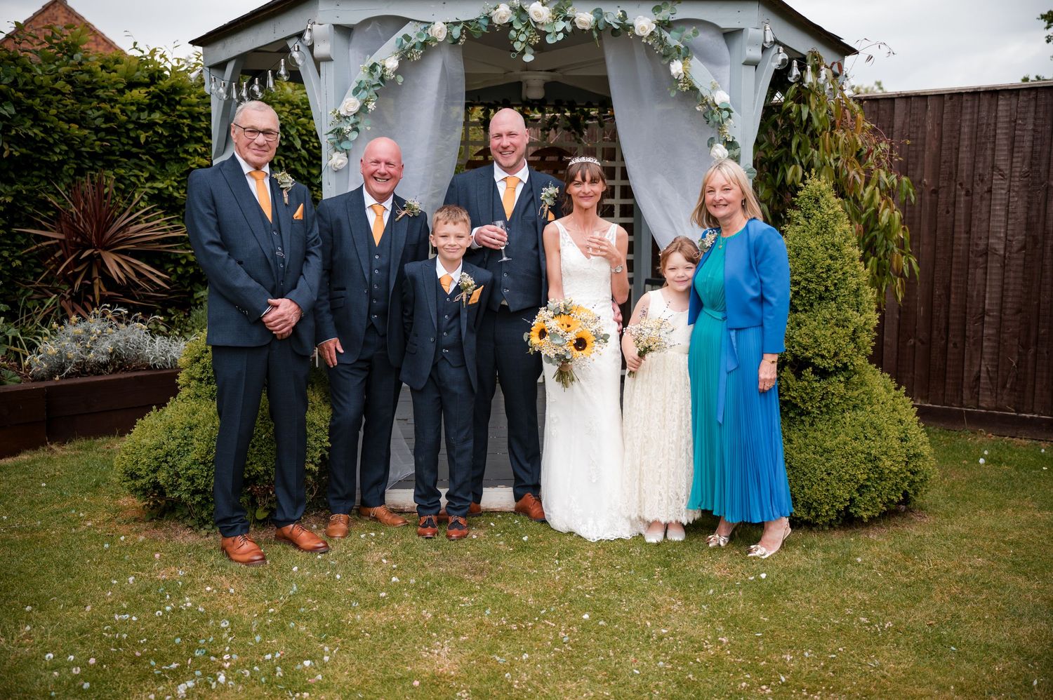 A group gathers under a white garden arch decorated with floral arrangements during an outdoor wedding ceremony.