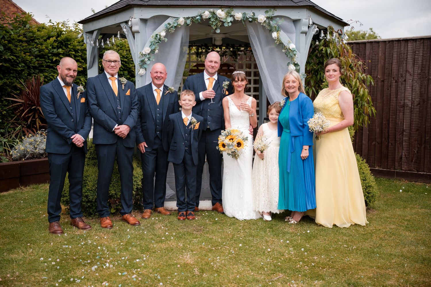 Wedding party members in formal attire pose together outdoors in front of a backyard garden setting.