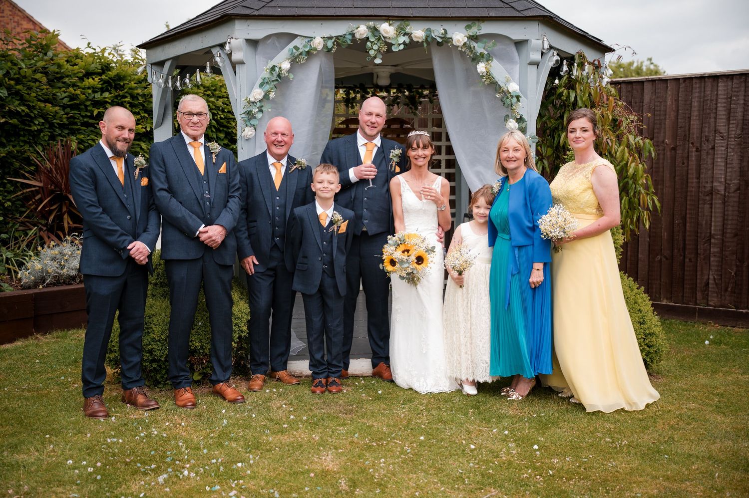 Wedding party poses together outdoors in formal attire with bridesmaids in yellow and blue dresses and groomsmen in navy suits.