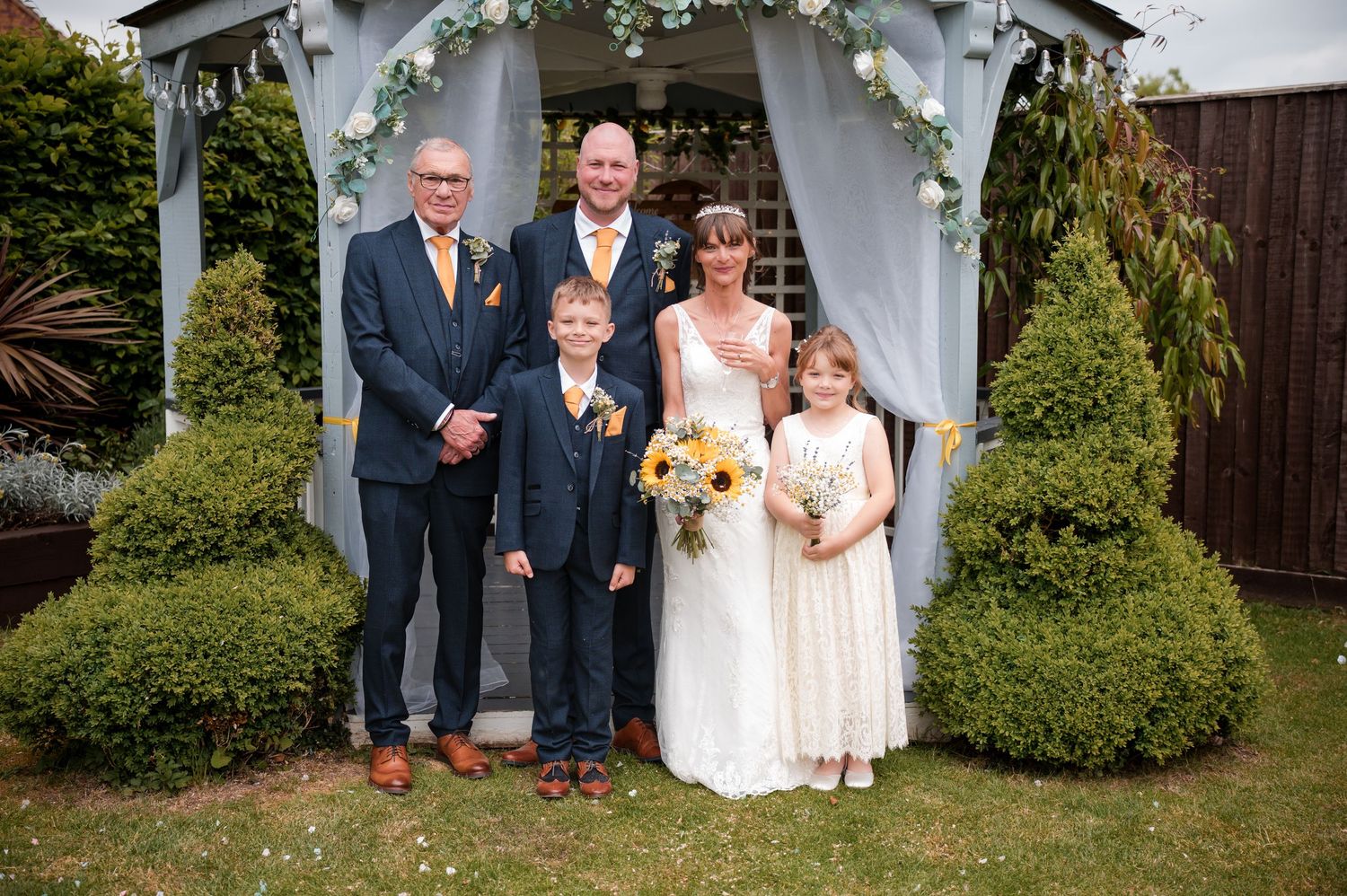A wedding party poses for photos in a lush garden setting with white gazebo and greenery in the background.