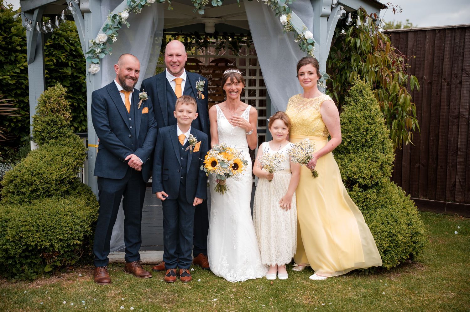 A wedding party in formal attire poses for photos outdoors in a garden setting with yellow and navy color scheme.
