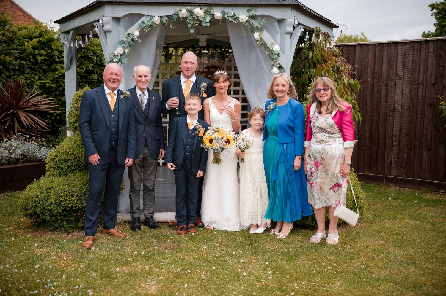 Wedding party members gather outdoors for group photos in a garden setting during a summer celebration.