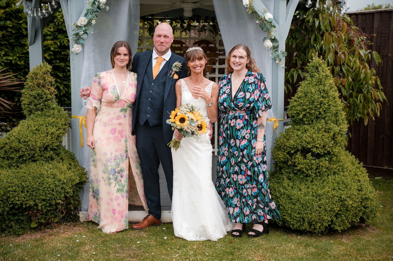 Four people pose together in formal attire under a white garden gazebo with greenery in the background.