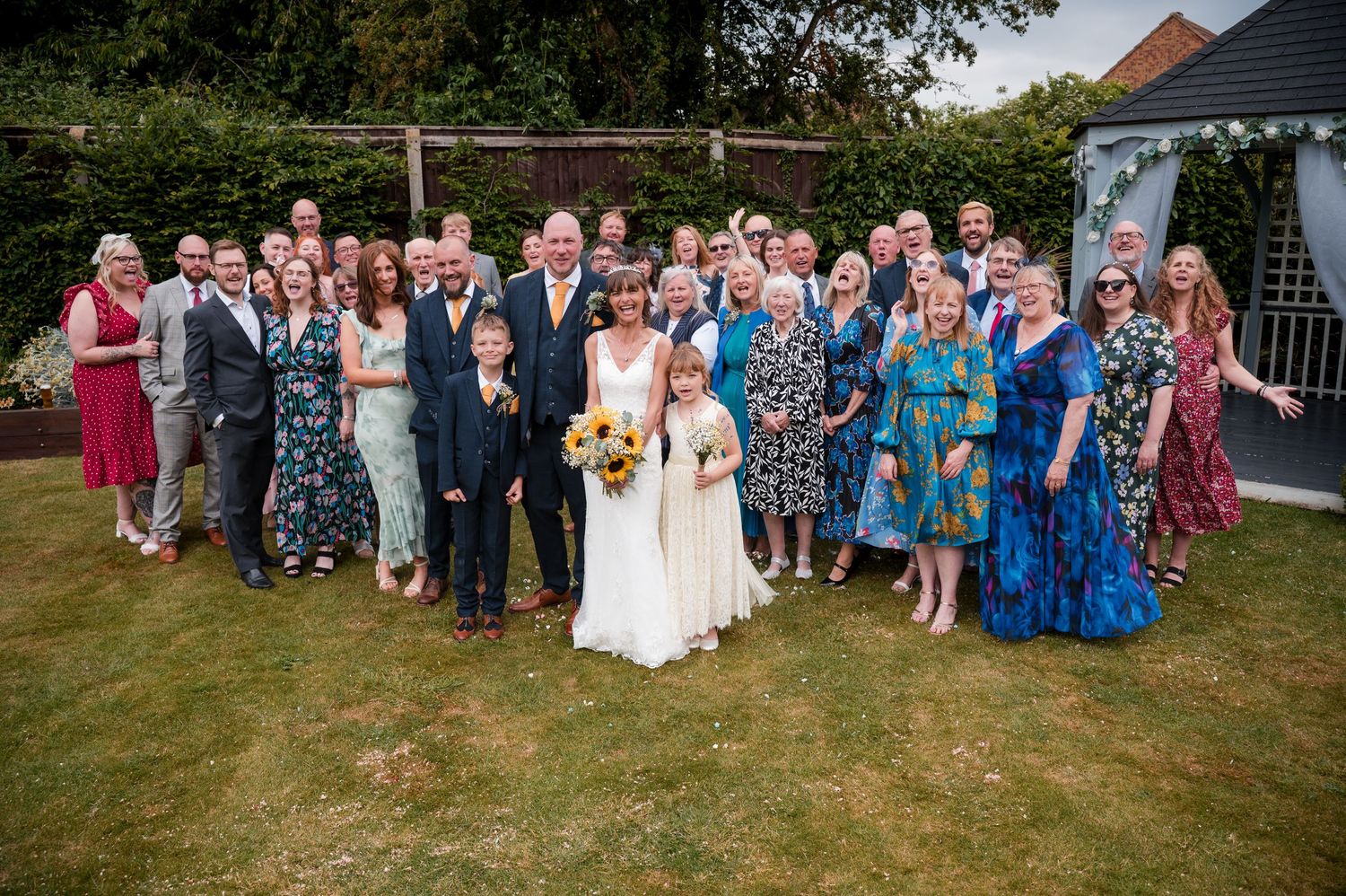 Large wedding party group poses together on a sunny lawn in front of a white gazebo.