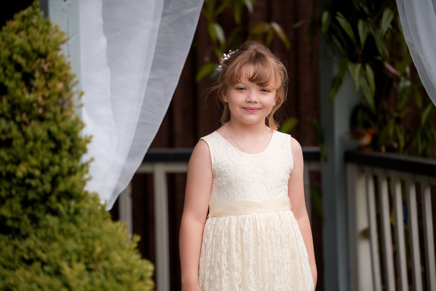 A child in a cream-colored sleeveless dress stands on a wooden porch with white curtains and greenery in background.