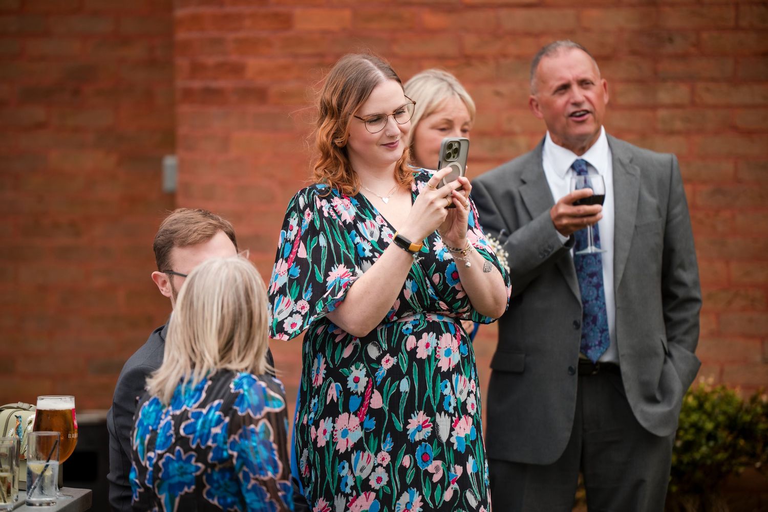 A group of wedding guests in floral dresses share laughter and conversation against a brick wall backdrop.