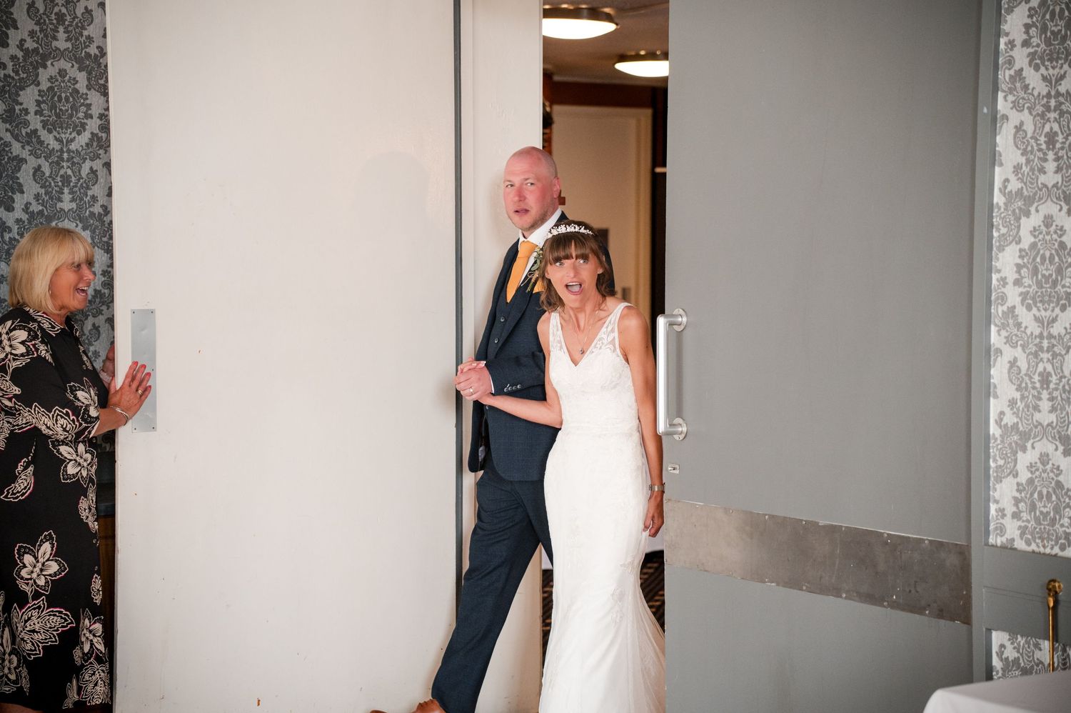 A bride in a white wedding dress shares a joyful moment with a companion in a hallway.