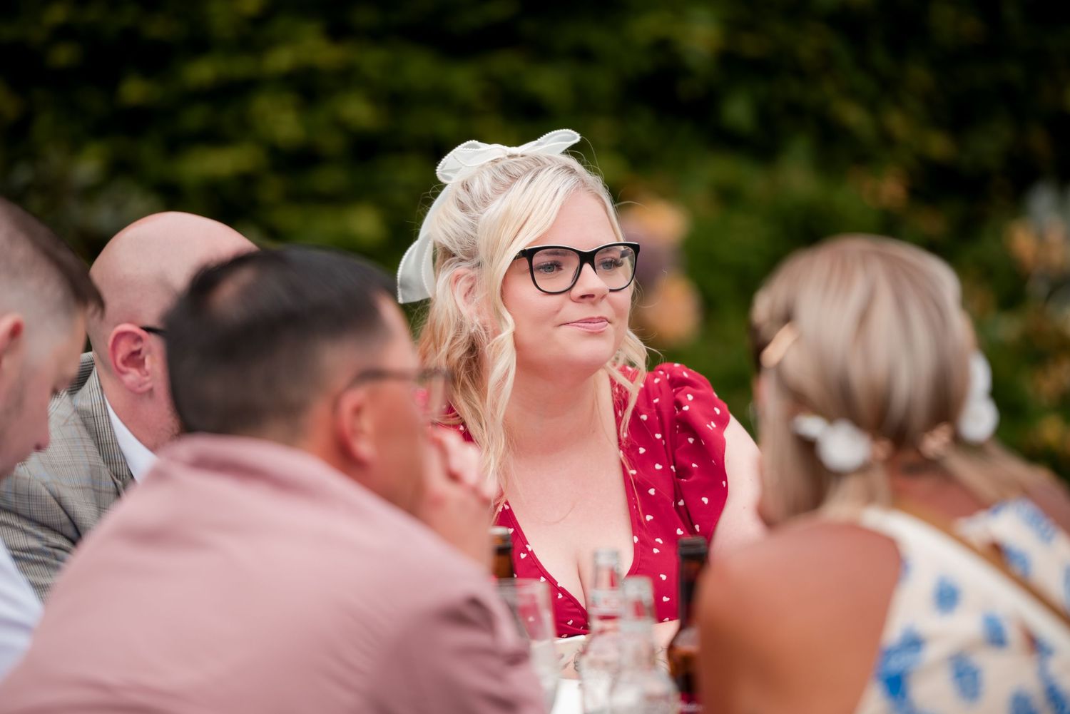 A wedding guest in a red dress and glasses smiles while seated at an outdoor reception table.