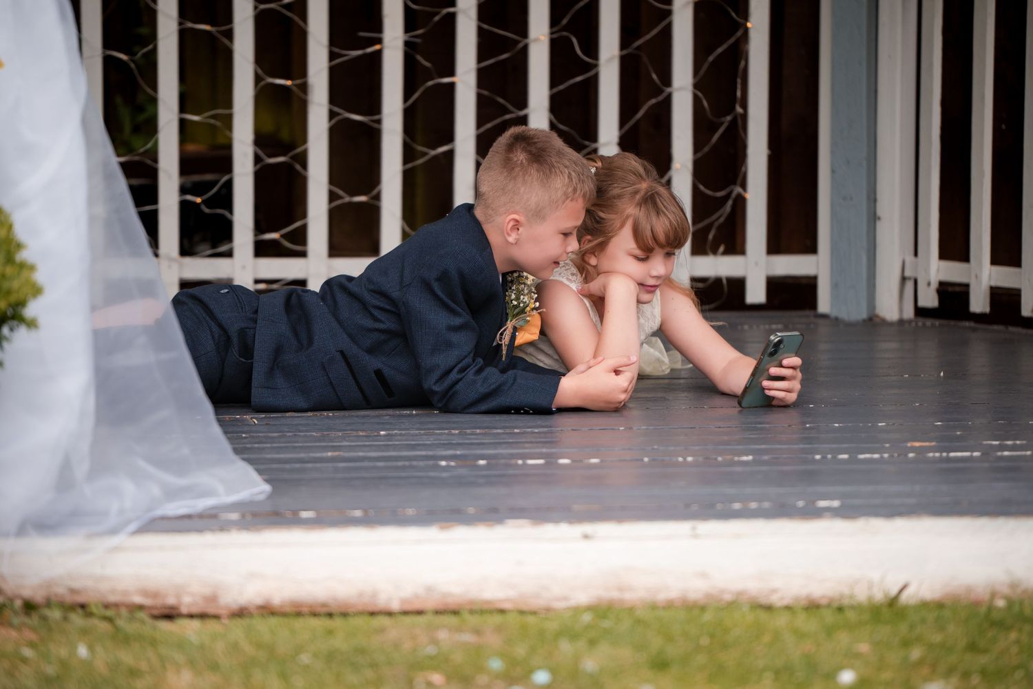 Wedding couple lays on porch floor while their dog peeks through railing bars in playful outdoor portrait.