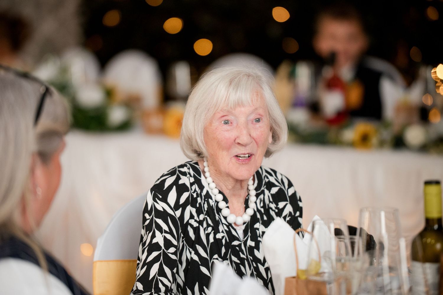 A group of guests enjoying a festive dinner party with glowing lights and table decorations in the background.