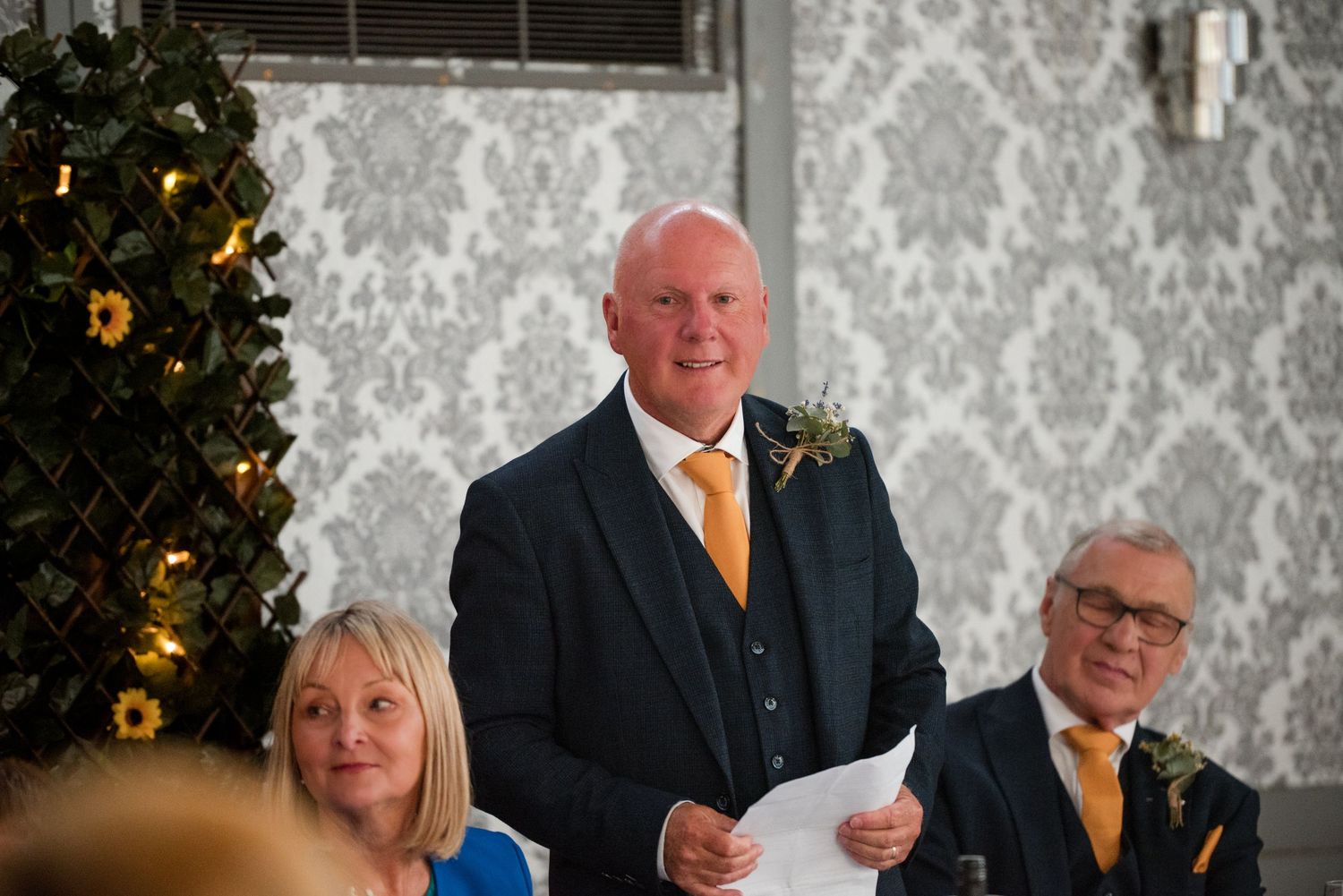 Wedding reception speeches being delivered in an elegant venue with decorative damask wallpaper and holiday decor.