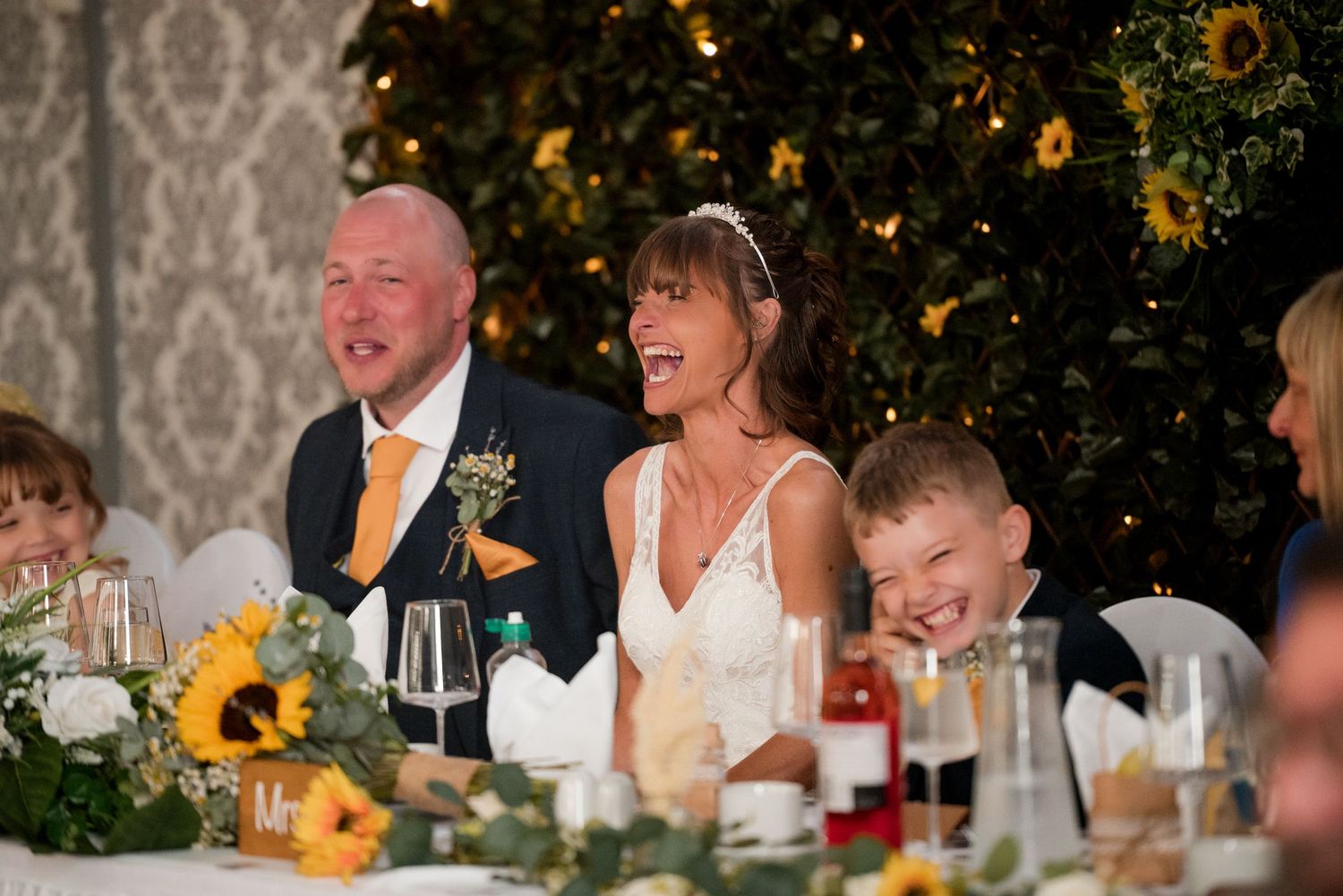 Wedding guests laugh and smile during dinner reception with sunflower centerpieces and candlelit tables.